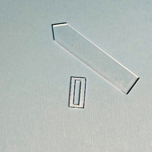 Könnte beinhalten: Zwei durchsichtige Plastikteile, eines ist ein langes Rechteck mit einer spitzen Endseite, das andere ist ein kleineres Rechteck mit einem Loch in der Mitte.