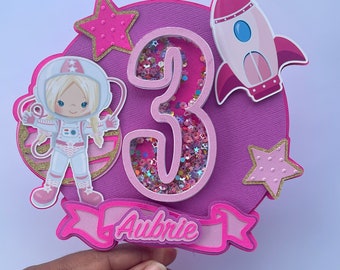 May include: Pink and gold astronaut cake topper with a 3D number 3, a rocket, stars, and the name Aubrie.