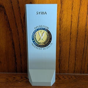 May include: A light gray vertical display plaque featuring the text SYRIA at the top and a circular emblem for Operation Combined Joint Task Force Inherent Resolve in the center, which includes two crossed swords and a gold laurel wreath.