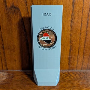 May include: A light gray, rectangular 3D-printed display stand featuring the embossed text IRAQ and IRAQI FREEDOM. Centered on the stand is a circular commemorative coin displaying a map of Iraq with the national flag and the text OPERATION IRAQI FREEDOM.
