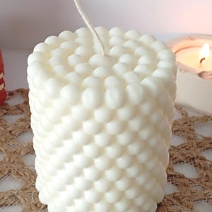 Peut inclure: Une bougie blanche cylindrique avec un motif texturé en forme de bulles. La bougie a une seule mèche et est posée sur une surface tissée de couleur beige. Une bougie allumée est visible en arrière-plan.