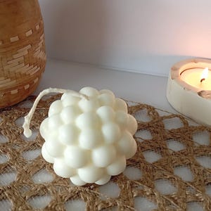 Peut inclure: Une bougie blanche en forme de bulles avec une mèche, ressemblant à un groupe de sphères. La bougie est posée sur une surface brune texturée, avec un panier tressé et une bougie allumée dans un support en béton en arrière-plan.