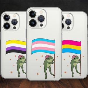 May include: Clear phone cases with illustrations of frogs holding flags representing non-binary, transgender, and pansexual identities. Each frog wears a pink hat and stands against a backdrop of small stars. The flags feature yellow, purple, black, pink, blue, and yellow.