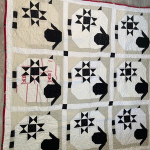 May include: A white and black quilt with a geometric pattern. The quilt is made up of nine squares, each with a different design. The squares are arranged in a 3x3 grid. The quilt is edged with a red border.