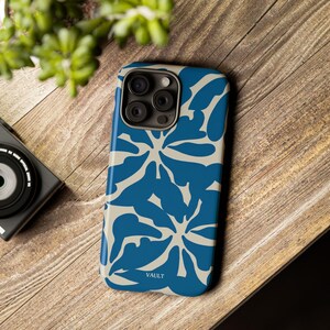 Può includere: Custodia per telefono con motivo floreale blu e beige con la scritta "VAULT" in basso. La custodia è su una superficie di legno con una fotocamera e vegetazione sullo sfondo.