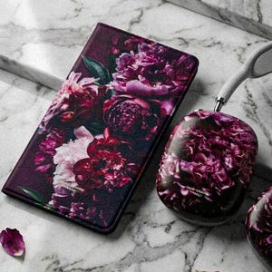 Può includere: Una custodia per telefono e cuffie con un design floreale di peonie rosa e viola. La custodia è rettangolare e le cuffie ovali. Sono su una superficie di marmo con petali sparsi.