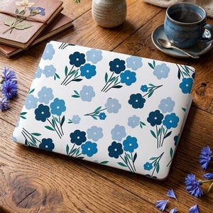 Può includere: Custodia per laptop bianca con motivo floreale blu. Il design presenta fiori blu stilizzati con centri blu scuro e steli verdi. Su un tavolo di legno, con un vaso di fiori blu e una tazza di tè.