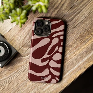 Può includere: Custodia per telefono con motivo bordeaux e beige con la scritta "VAULT" stampata in basso. La custodia è su una superficie di legno, con una fotocamera e del verde sullo sfondo.