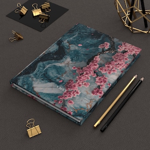 Può includere: Un diario con un design in marmo verde acqua e fiori di ciliegio rosa. La parola "VAULT" è stampata sulla copertina. Matite dorate e nere, insieme a fermagli dorati, sono disposte attorno al diario su una superficie grigia.
