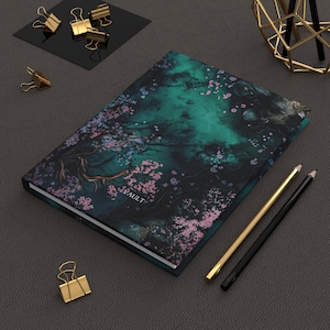 Può includere: Diario con copertina rigida con una copertina verde scuro e nera con un motivo floreale con fiori rosa e un ramo d'albero. La parola "VAULT" è stampata in basso a sinistra. Sono visibili anche matite dorate e nere e fermagli dorati.
