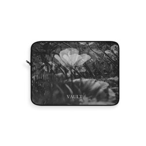 Può includere: Custodia per laptop in bianco e nero con un design floreale. L'immagine mostra fiori in un campo, con la parola "VAULT" stampata in basso. La custodia ha una cerniera nera.