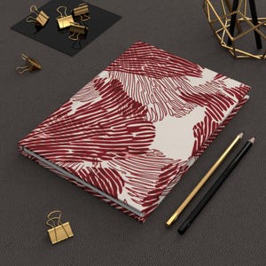 Può includere: Un diario con copertina rigida con sfondo color crema e un motivo ripetuto di disegni floreali astratti rossi. Matite dorate e nere e fermagli dorati sono disposti attorno al diario.