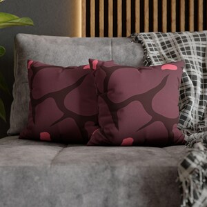 Può includere: Due cuscini quadrati con sfondo bordeaux e un motivo astratto marrone scuro. I cuscini hanno accenti rosa e sono posizionati su un divano grigio. Una coperta fantasia è visibile sullo sfondo.