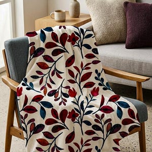 May include: A cream-coloured throw blanket with a botanical design of red, blue, and purple leaves and flowers on black stems, draped over a chair. The blanket is on a wooden chair with a grey seat. A small succulent plant in a concrete pot is nearby.