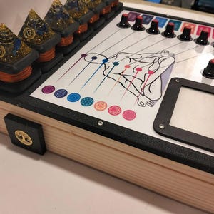 May include: A wooden box with a white top featuring a diagram of a human figure with chakra symbols. It has black knobs, a square frame, and several pyramid-shaped objects with copper wire and crystals.