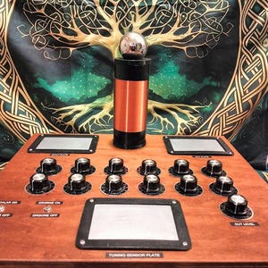 May include: A wooden device with multiple knobs, switches, and a central cylindrical component wrapped in copper wire. A silver sphere sits atop the cylinder. The text "TUNING SENSOR PLATE" is visible on the device. The background features a tapestry with a tree design.