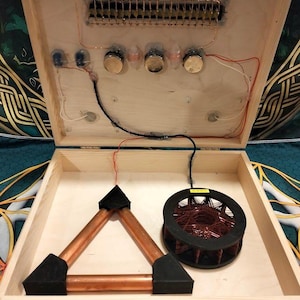 May include: A light-colored wooden box containing electronic components. The lid features a circuit board with copper wiring, three silver knobs, and a resistor array. Inside the base sits a copper pipe triangle and a circular black coil spool.