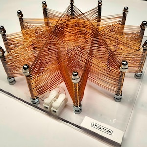 Può includere: Un assemblaggio di bobine di filo di rame a forma di stella montato su una base in acrilico trasparente. Il design presenta molteplici montanti metallici verticali con tappi color argento, interconnessi da sottile filo di rame. Un'etichetta bianca riporta il testo LK.23.11.25.