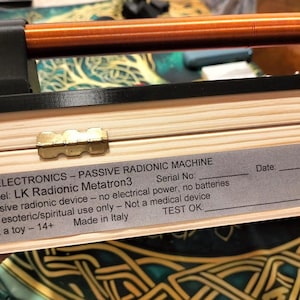 May include: A light-colored wooden box with a black top and a copper-colored rod, featuring a silver label that reads: LK ELECTRONICS - PASSIVE RADIONIC MACHINE, Model: LK Radionic Metatron3, Passive radionic device, Made in Italy, Not a toy - 14+.
