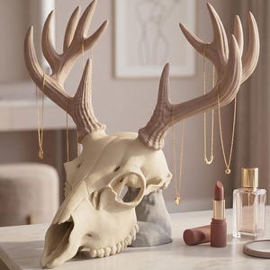 May include: A cream-coloured deer skull sculpture with large antlers, used as a jewellery holder. Gold necklaces hang from the antlers. Lipstick, perfume, and a compact mirror sit on the marble surface in front of the skull.