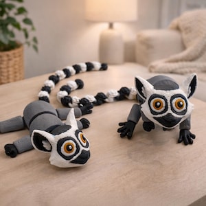 Peut inclure: Deux figurines de lémuriens sur une table en bois. Les lémuriens sont gris et blancs avec des détails noirs et de grands yeux orange. L'un des lémuriens est allongé et l'autre est debout. Une queue rayée noire et blanche relie les deux.