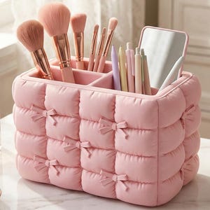 Puede incluir: Un organizador de maquillaje acolchado rosa con múltiples compartimentos, que contiene varios pinceles de maquillaje, bolígrafos y un pequeño espejo. El organizador tiene un detalle de lazo en cada sección acolchada. Los pinceles son rosas y dorados.