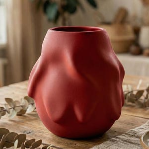 May include: A matte red ceramic vase with an organic, textured design. The vase has a unique, sculptural shape and sits on a rustic wooden table. The background includes a neutral-toned dining setting with natural light.