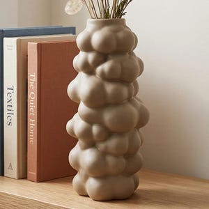 May include: A beige, cloud-shaped ceramic vase filled with dried flowers and grasses. The vase sits on a light wood shelf next to three books with titles including "Forms" and "Textiles". The arrangement is set against a neutral wall.
