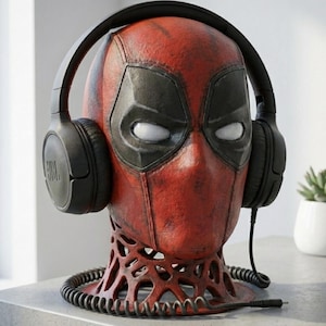 May include: A red and black Deadpool head bust with black headphones. The bust has a detailed design, with a textured appearance. The headphones are black and have the JBL logo. The bust is on a decorative base.