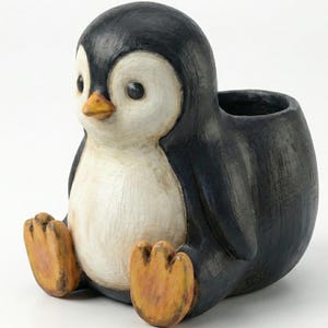 Puede incluir: Una maceta de cerámica con forma de pingüino, con cuerpo negro, pecho blanco y patas y pico naranjas. El pingüino está sentado con las piernas extendidas y tiene la espalda ahuecada para plantar pequeñas plantas o guardar objetos. La maceta tiene un acabado envejecido.