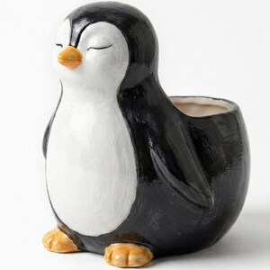 May include: A ceramic penguin planter in black, white, and orange. The penguin has closed eyes and a small opening at the top for plants. The planter is a decorative item for home or office.