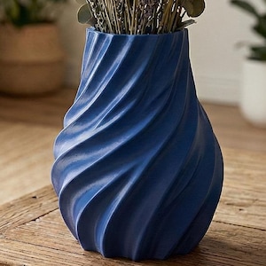 May include: A blue vase with a spiral design holds a bouquet of dried lavender and eucalyptus. The vase is made of a solid material and sits on a wooden table. The background includes plants and a neutral-toned curtain.