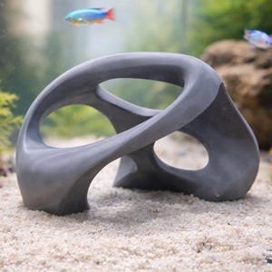 Op de afbeelding: Een grijze, abstracte aquariumdecoratie met een gebogen, open ontwerp. De sculptuur staat op een zandbed in een aquarium, omringd door planten en kleurrijke vissen. De tank is gevuld met water.