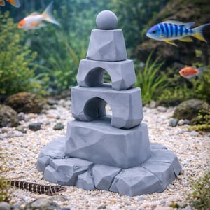 Puede incluir: Decoración gris apilada para acuario, con forma de torre de piedra con una bola en la parte superior. La torre tiene arcos y se asienta sobre una base rocosa. Varios peces nadan en el fondo.