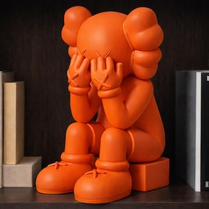 May include: Orange vinyl art figure in a seated pose, hands covering the eyes. The figure has large, rounded ears and oversized feet. The hands and feet have stitched details. The figure is sitting on a small orange block.