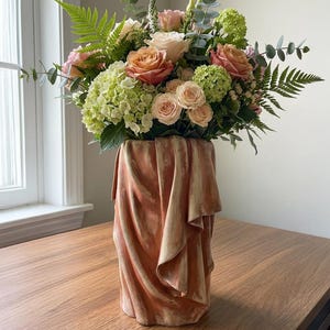 May include: A floral arrangement featuring peach and pink roses, green hydrangeas, and ferns in a draped terracotta vase. The vase has a textured, draped design. The arrangement sits on a wooden table near a window.