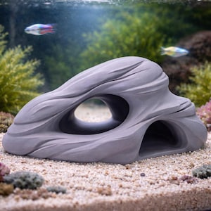 Op de afbeelding: Grijze, rotsvormige aquariumdecoratie met een grote centrale opening en kleinere openingen eronder. Het gestructureerde oppervlak bootst natuursteen na en biedt vissen een schuilplaats. In een aquarium met zand en planten.