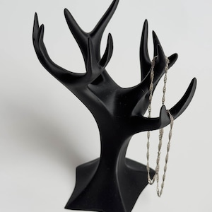 Puede incluir: Un expositor de joyas negro con forma de árbol y múltiples ramas. Un collar de cadena plateada cuelga de una de las ramas. El expositor de joyas está hecho de un material sólido y mate y está sobre un fondo blanco.