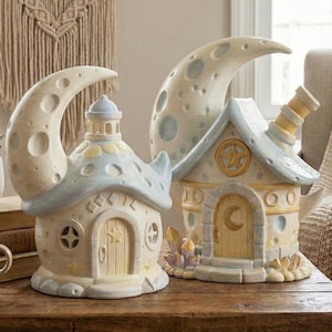 Pode incluir: Duas casas de fadas de cerâmica creme e caprichosas com detalhes de lua crescente. Uma tem uma pequena torre, a outra uma chaminé. Ambas têm telhados azuis claros e detalhes de porta de estrela e lua. Cristais decorativos na base de uma casa.
