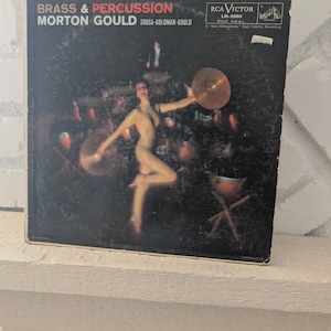 May include: Vintage vinyl record album cover for "Brass & Percussion" by Morton Gould. The cover features a nude woman playing a cymbal, with the album title and artist's name in white text. The RCA Victor logo is visible.