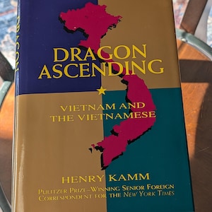 Puede incluir: Un libro de tapa dura titulado "Dragon Ascending" con una cubierta dorada, verde azulado y azul marino. El libro presenta un mapa de Vietnam en letras rojas y doradas. También son visibles el nombre del autor, Henry Kamm, y el subtítulo "Vietnam y los vietnamitas".