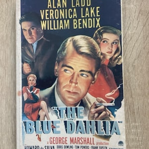 Puede incluir: Cartel de película vintage de "The Blue Dahlia" con Alan Ladd, Veronica Lake y William Bendix. El cartel tiene un fondo azul oscuro con imágenes de los actores y el título de la película.