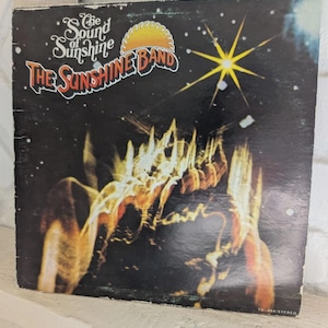 Puede incluir: Funda de álbum de vinilo vintage para "The Sound of Sunshine" de The Sunshine Band. La portada presenta un fondo oscuro con una estrella amarilla y llamas abstractas naranjas y amarillas. El nombre de la banda está en texto naranja y blanco.