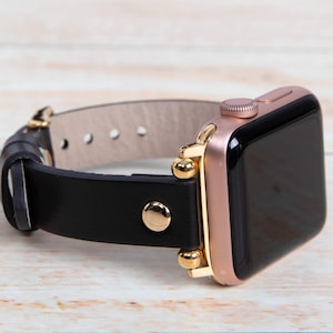 May include: A rose gold smartwatch with a black screen and a black leather strap. The strap has gold-coloured hardware and a light tan interior. The watch is displayed on a light wood surface.