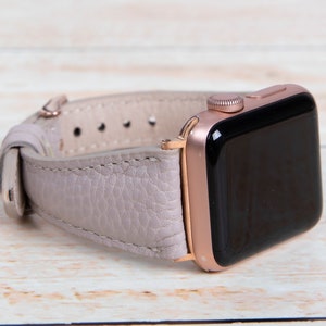 May include: A rose gold Apple Watch with a pale purple leather strap. The watch face is black, and the strap is secured with a rose gold buckle. The watch is on a light wood surface.