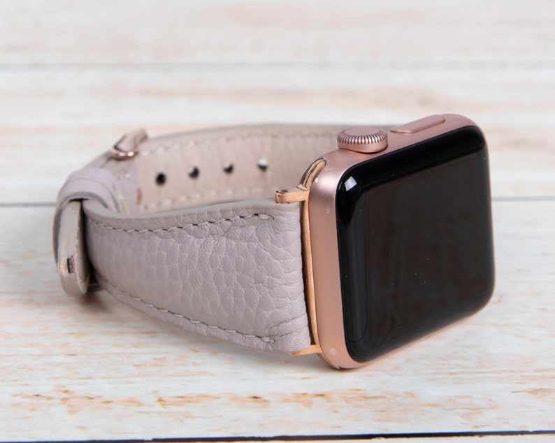 May include: A rose gold Apple Watch with a pale purple leather strap. The watch face is black, and the strap is secured with a rose gold buckle. The watch is on a light wood surface.