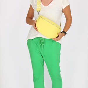 May include: A woman wearing a white t-shirt with a pink trim and green pants is holding a yellow fanny pack with a white and yellow patterned strap.