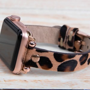 May include: A rose gold Apple Watch with a leopard print leather band. The band has gold studs and a rose gold buckle.