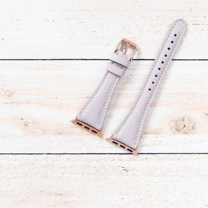 May include: A light purple leather watch strap with a textured finish. The strap features rose gold-coloured metal clasps and connectors. The watch strap is displayed on a light wood grain surface.