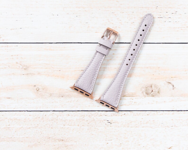 May include: A light purple leather watch strap with a textured finish. The strap features rose gold-coloured metal clasps and connectors. The watch strap is displayed on a light wood grain surface.